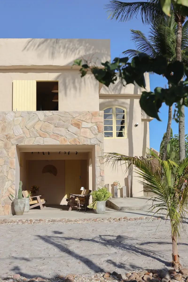 Fachada de cal e pedra com janelas amarelas, entre coqueiros